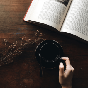 A hand holding a black cup of tea on a wooden table next to an open book and a sprig of herbs.