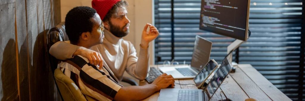 Two people in front of a computer screen with code. One is white, is wearing a red beanie and has a beard. One is black, and is wearing glasses.