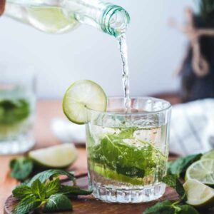 A bottle pouring water into a glass with a lime garnish and mint leaves. The glass is on a wooden board surrounded by lime slices and mint leaves.