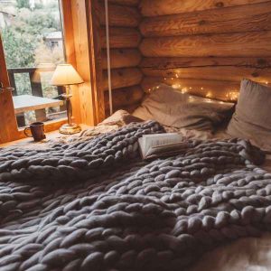 A photo in a cozy log cabin. There is a bed with a heavy textured grey knit blanket, and some soft lighting by a window.